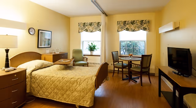 A well-lit senior living bedroom with a single bed covered in a beige quilt, a bedside table with a lamp, a green armchair, a small round dining table with two chairs, two windows with floral valances, a flat-screen TV on a stand, and light yellow walls.