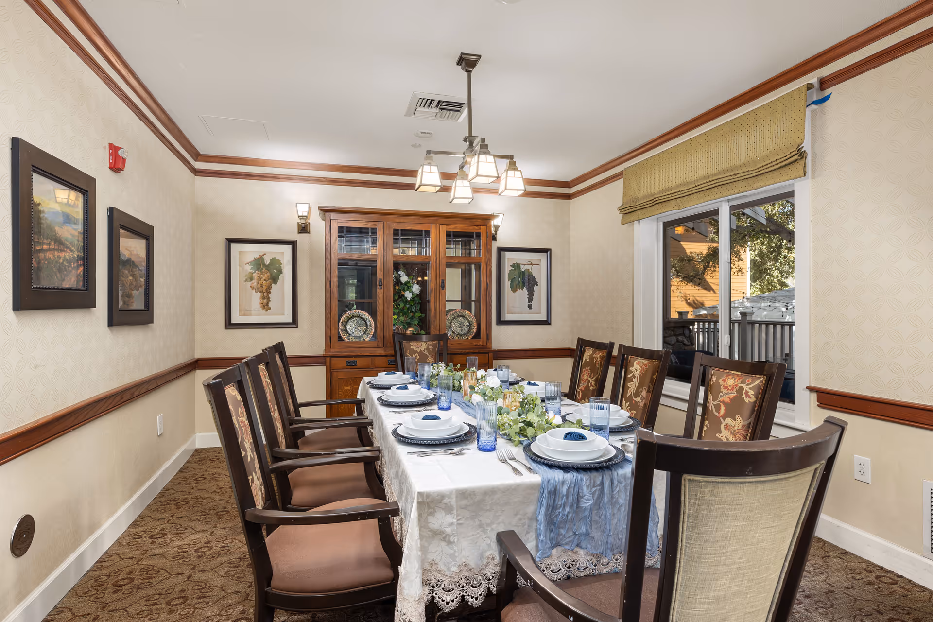 A formal dining room with a long rectangular table set for eight people. The table is covered with a white lace tablecloth and a blue runner, and is set with plates, bowls, glasses, and silverware. There are floral centerpieces along the middle of the table. The room has beige walls with wooden trim, framed artwork on the walls, a wooden china cabinet at the far end, and a window with a beige roman shade letting in natural light.