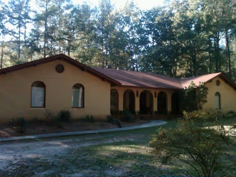 Exterior view of a single-story building with a brown roof and beige walls surrounded by trees and greenery. The building has arched windows and a covered porch area with arches.