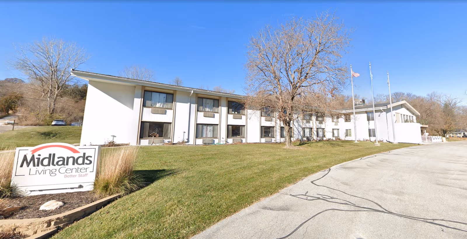 Exterior view of Midlands Living Center, a two-story white building with multiple windows, a grassy lawn, a few leafless trees, and three flagpoles with flags. A sign in front reads 'Midlands Living Center Better Staff'. The sky is clear and blue.