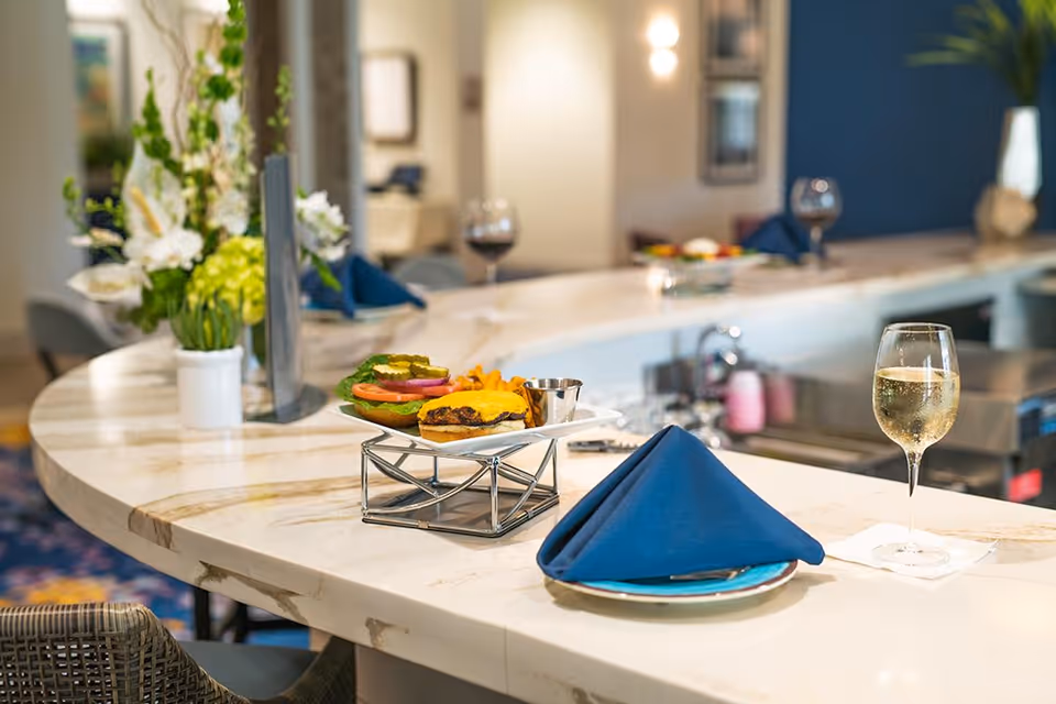 A curved marble countertop with a plate of cheeseburger and fries, a glass of white wine, and a blue folded napkin on a plate. In the background, there are more glasses of wine and a vase with white and green flowers, suggesting a dining or bar area in a senior living facility.