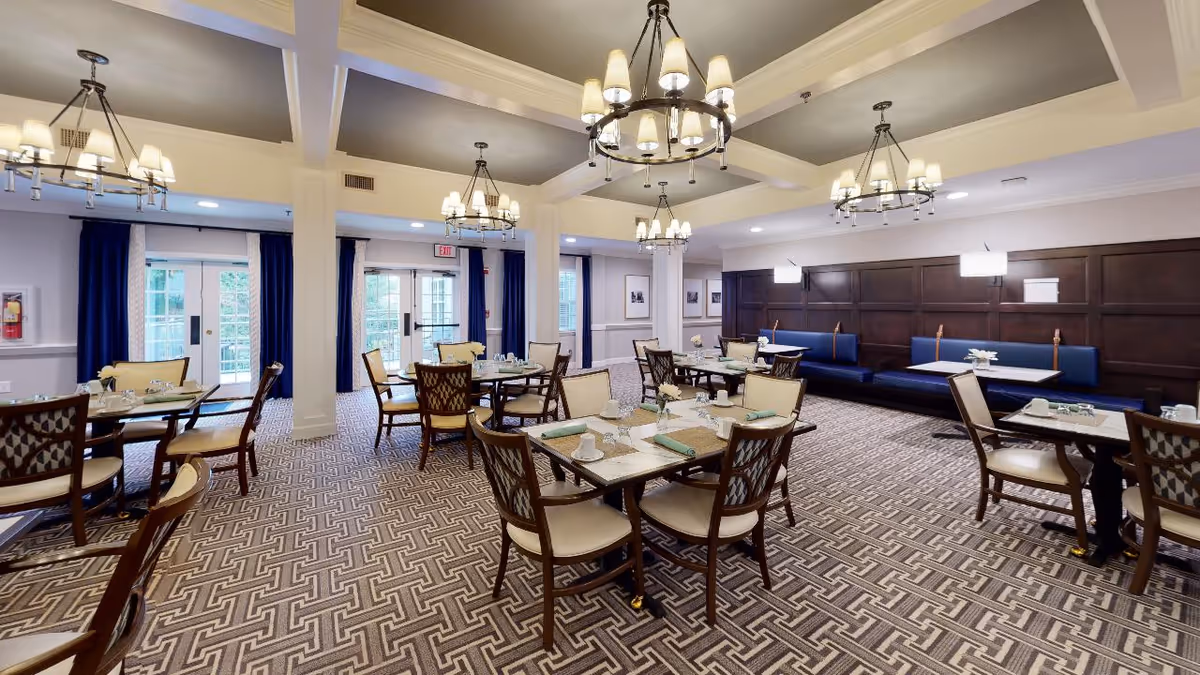 A spacious dining room with multiple wooden tables and chairs arranged neatly. The room features patterned carpet flooring, dark wood paneling on one wall with blue cushioned bench seating, and large windows with blue curtains allowing natural light. Several chandeliers hang from the ceiling providing warm lighting.