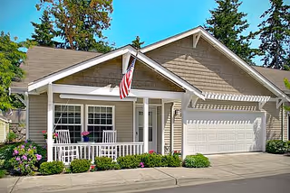 Exterior view of a single-story residential cottage with beige siding, a white front porch with rocking chairs, an American flag mounted on the porch, a two-car garage, and surrounding greenery including bushes and trees.