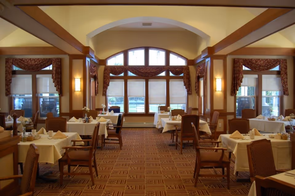 A spacious dining room with multiple tables covered in white tablecloths, each set with napkins, plates, and utensils. The room features large windows with decorative valances, wooden beams on the ceiling, and warm lighting from wall sconces. Chairs with armrests are arranged around the tables, and the carpet has a patterned design.