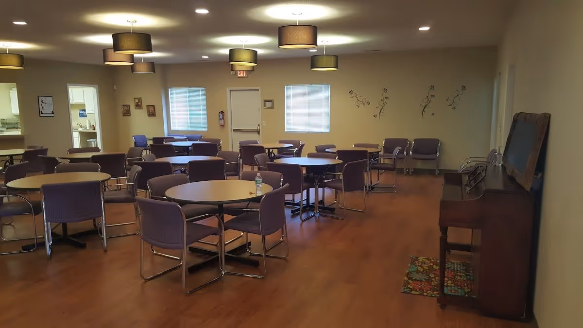 A spacious room with multiple round tables and purple chairs arranged for group seating. The room has wooden flooring, several ceiling lights with brown lampshades, two windows with blinds, and a piano against the right wall with a colorful rug underneath. There are some framed pictures on the walls and a fire extinguisher near the exit door.