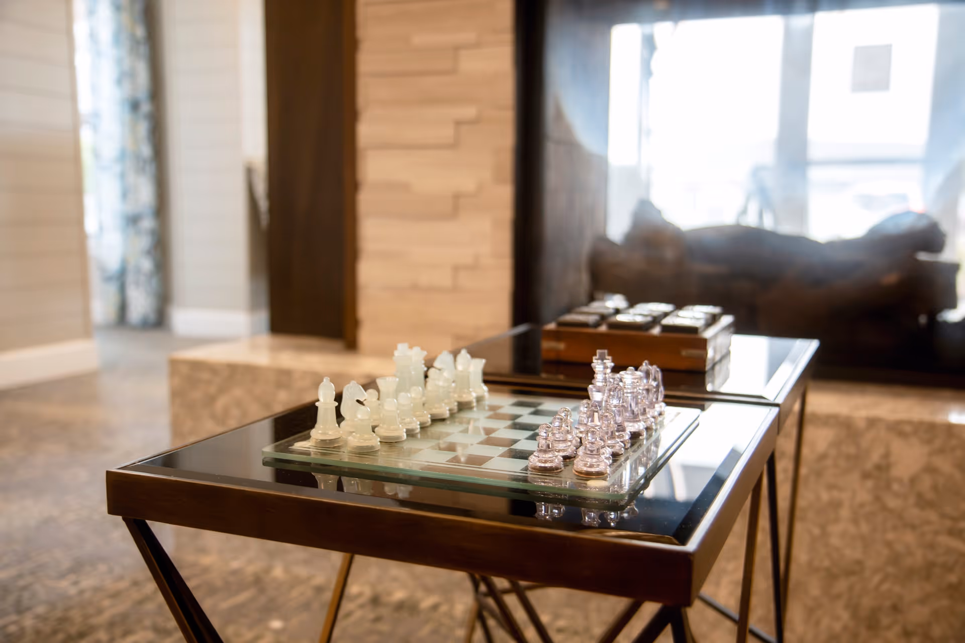 A glass chess set arranged on a glass chessboard placed on a dark wooden table in a cozy indoor setting with a stone fireplace and a window in the background.
