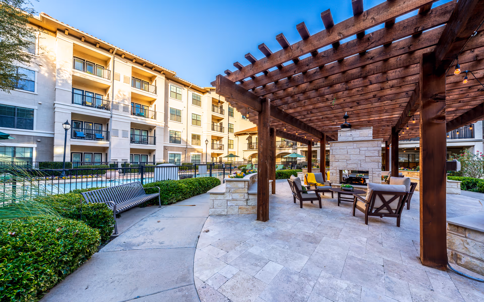Outdoor patio area at Conservatory At Plano featuring a wooden pergola with string lights, cushioned chairs arranged around a stone fireplace, a bench along a pathway, and a multi-story residential building in the background under a clear blue sky.