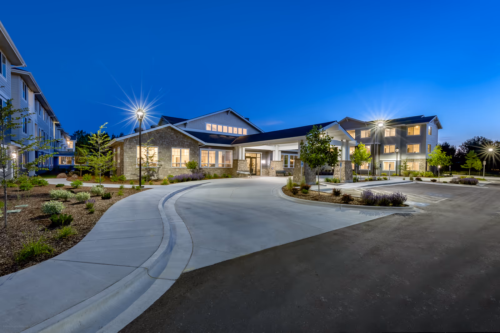 Exterior view of Revel Eagle senior living facility at dusk, showing a well-lit entrance with a covered drop-off area, landscaped surroundings, and multi-story residential buildings in the background under a clear blue sky.
