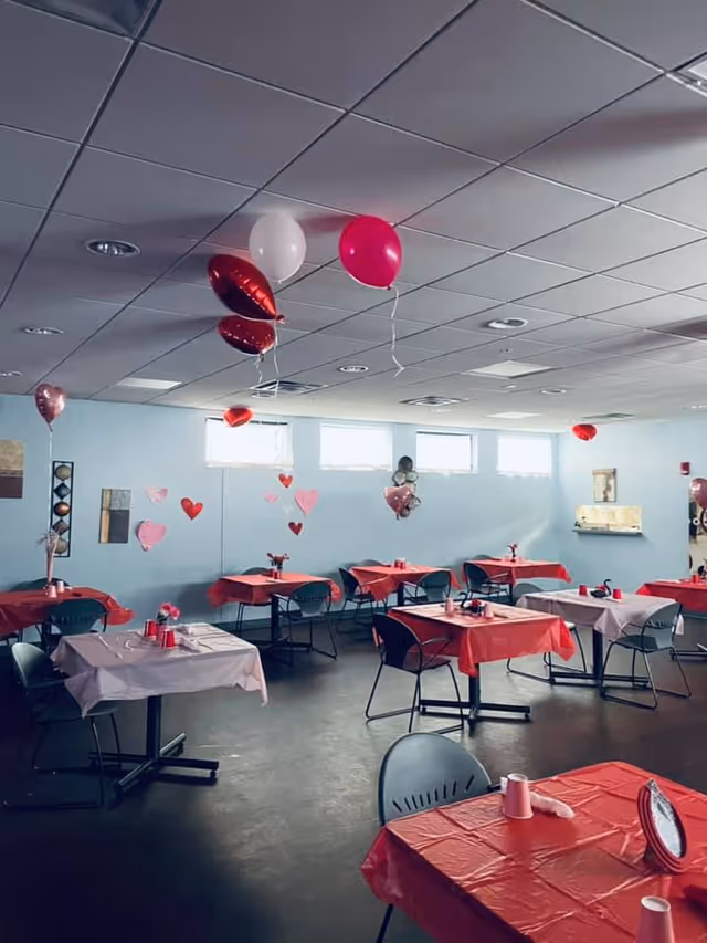 A dining room decorated with red and pink tablecloths, heart-shaped balloons, and paper hearts on the walls, suggesting a Valentine's Day theme. Several tables with chairs are arranged throughout the room, each table set with cups and small decorations. The room has a drop ceiling with fluorescent lights and windows letting in natural light.