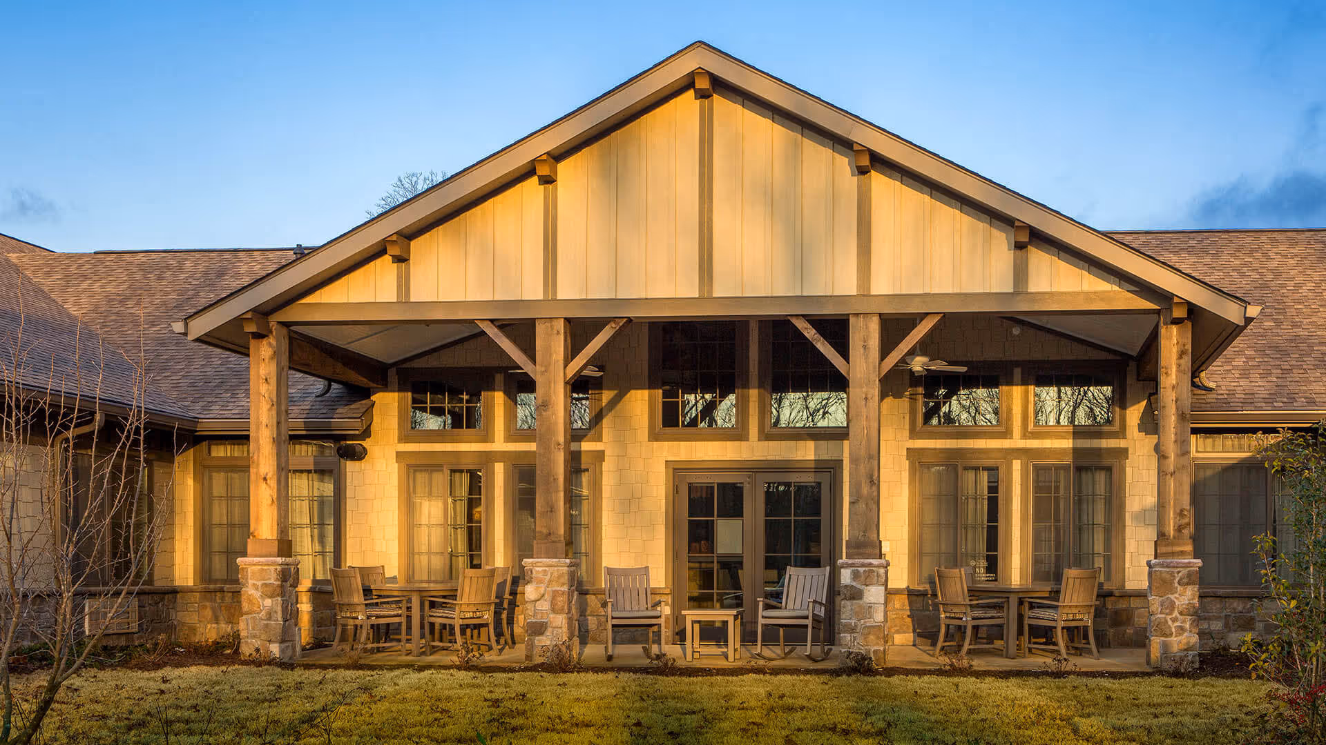 Exterior view of a senior living facility named Thrive at Brow Wood, featuring a covered patio area with wooden pillars, stone bases, and outdoor seating including tables and chairs. The building has large windows and a peaked roof under a clear blue sky.