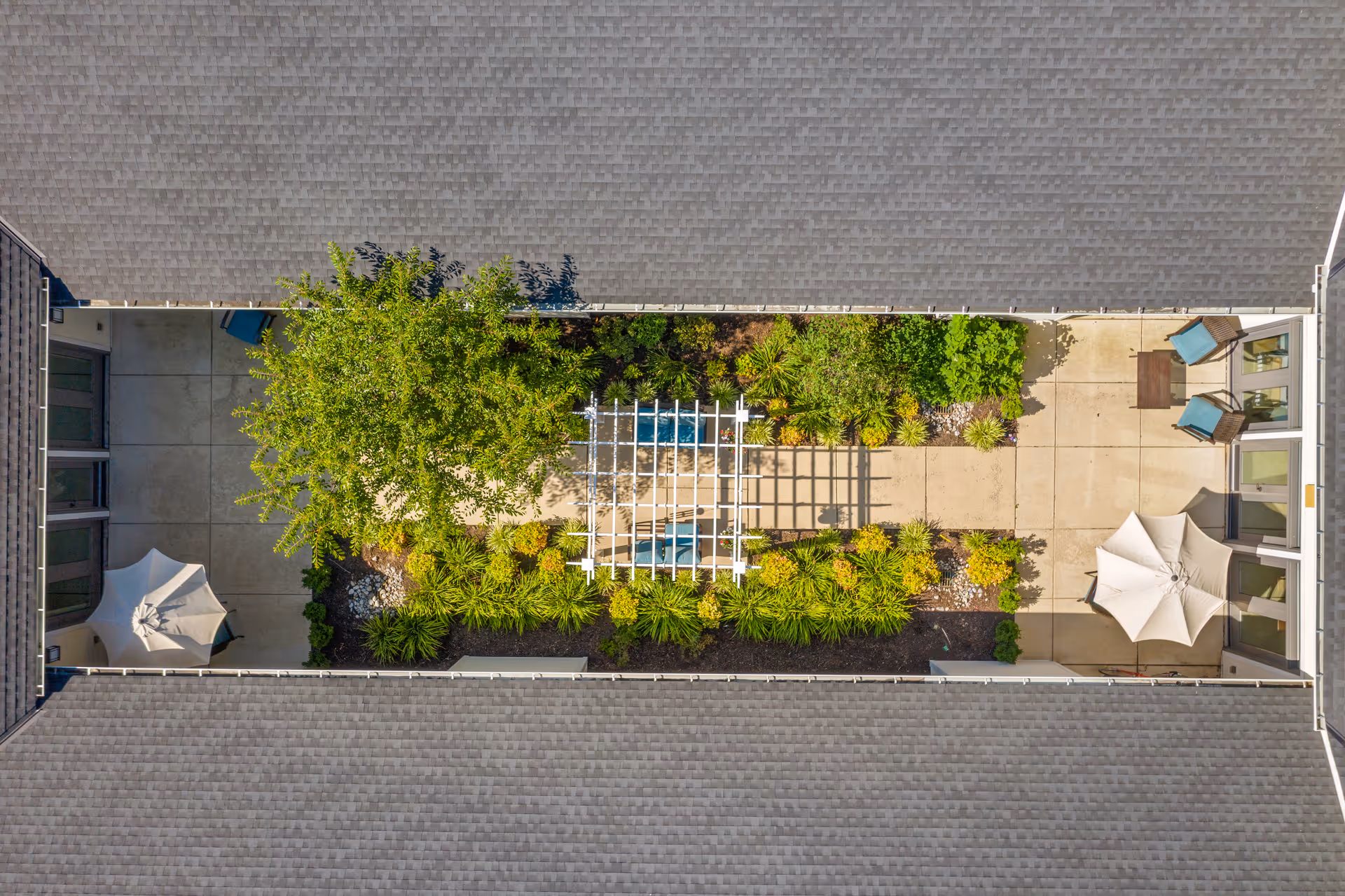 Aerial view of a rectangular courtyard between building roofs featuring a central pergola, planted beds, patio seating, and umbrellas.