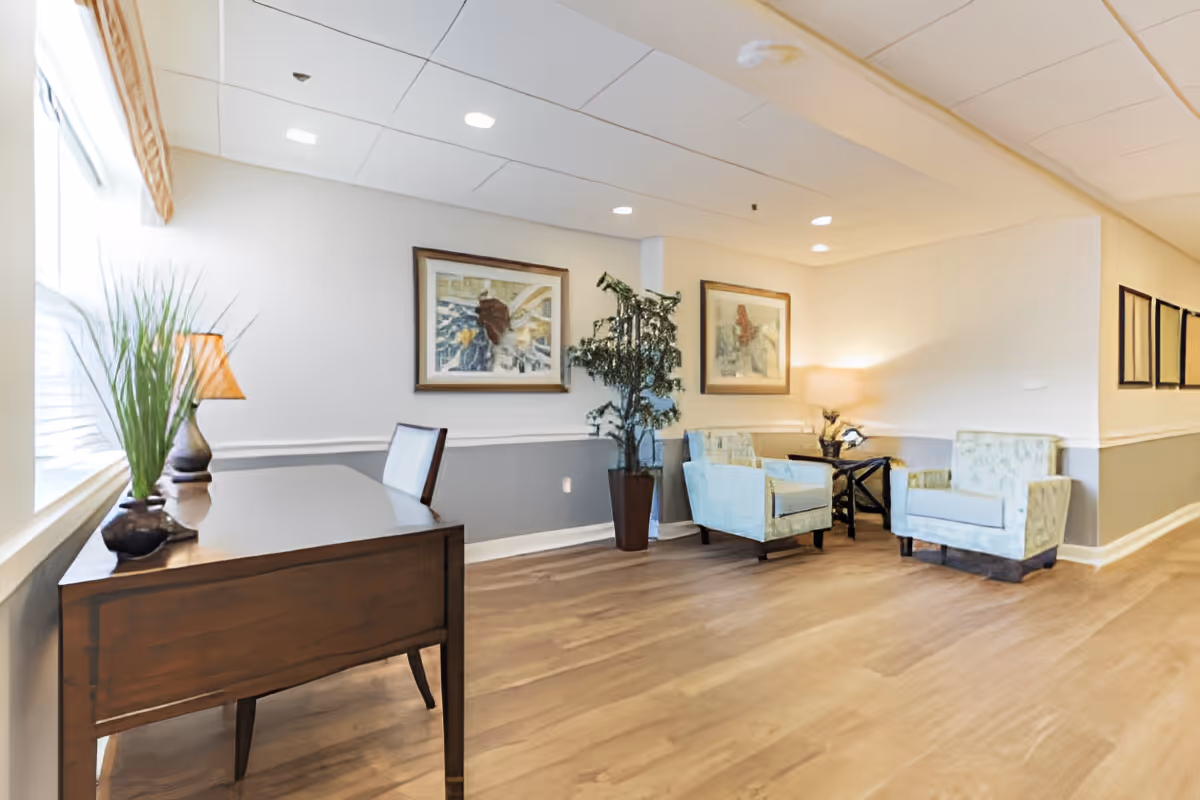 A bright and spacious interior seating area with two upholstered armchairs, a small side table with a lamp, framed artwork on the walls, a potted plant, and a wooden desk with a chair near a window with blinds.