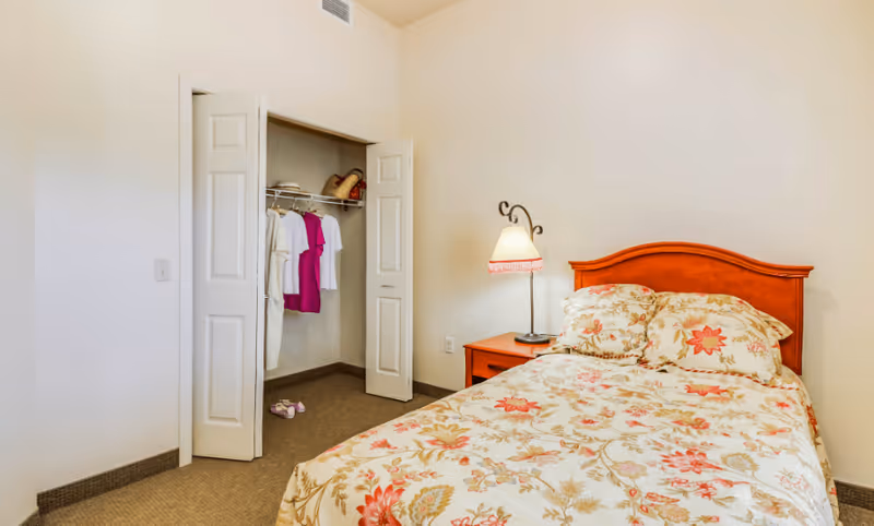 A bedroom with a floral patterned bedspread on a bed with a wooden headboard. Next to the bed is a wooden nightstand with a decorative lamp. An open closet with white bi-fold doors reveals hanging clothes and a few items on the shelf.