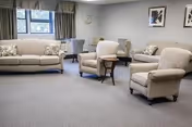 A senior living center common area with a beige sofa, two beige armchairs, a small wooden side table, and a desk with a chair near a window. The room has gray carpet, light gray walls, framed artwork, and a clock on the wall.