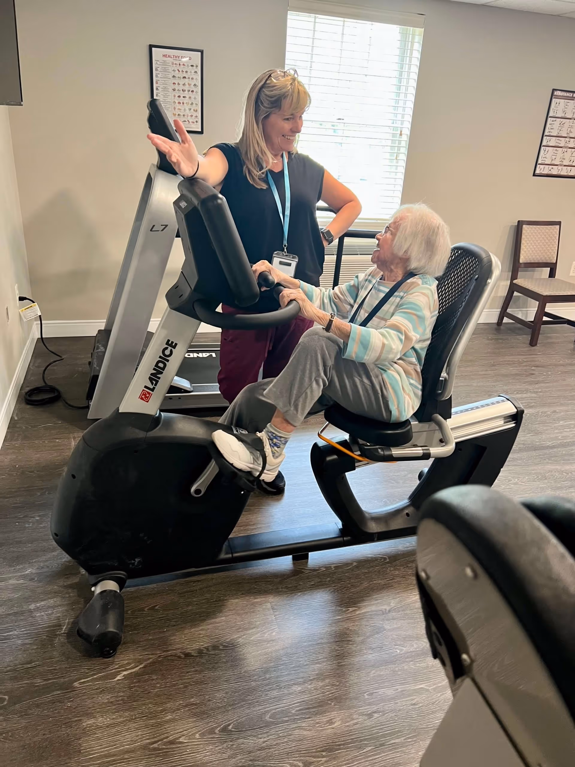 An elderly woman is seated on a recumbent exercise bike while a smiling female staff member stands beside her, engaging in conversation. The room has wooden flooring, a window with blinds, and exercise equipment in the background.