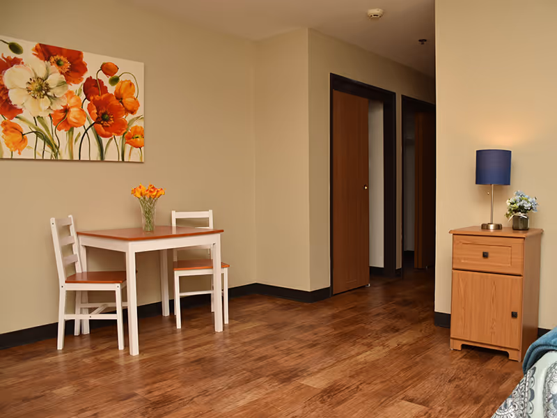 A small dining area in an assisted living facility with a wooden table and two white chairs. A vase with orange flowers is on the table. On the wall above the table is a painting of orange and white flowers. To the right, there is a wooden nightstand with a blue lamp and a small flower arrangement. The floor is wooden, and there are two open doorways in the background.