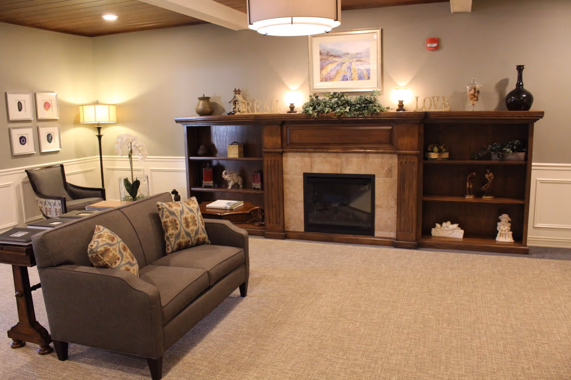 A cozy living room area in a senior living facility featuring a gray sofa with patterned cushions, a wooden side table, a floor lamp, and a chair. The room has a wooden fireplace mantel with decorative items and shelves on either side, a framed painting above the fireplace, and soft beige carpeting.