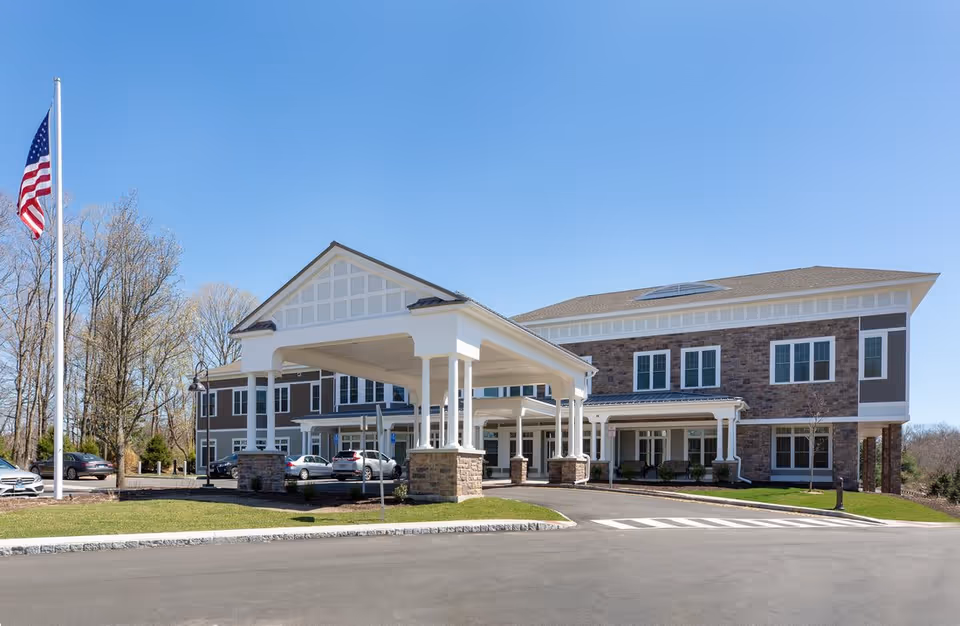 Exterior view of The Residence at Selleck's Woods building with a covered entrance, several parked cars, an American flag on a flagpole, and clear blue sky.