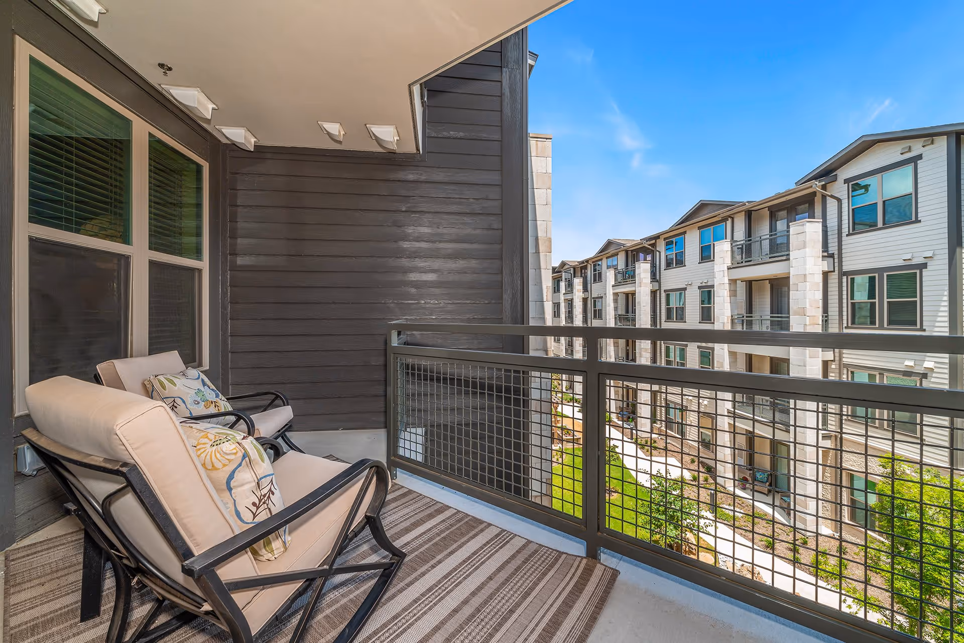 Small furnished balcony with two cushioned chairs overlooking an apartment courtyard and neighboring buildings.
