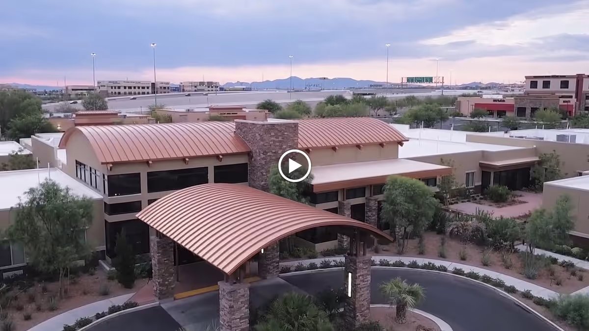 Aerial view of Canyon Vista Post-Acute facility showing a modern building with curved copper-colored roofs, stone pillars at the entrance, surrounding desert landscaping with trees and shrubs, and a circular driveway in front. The background includes distant mountains and a highway.