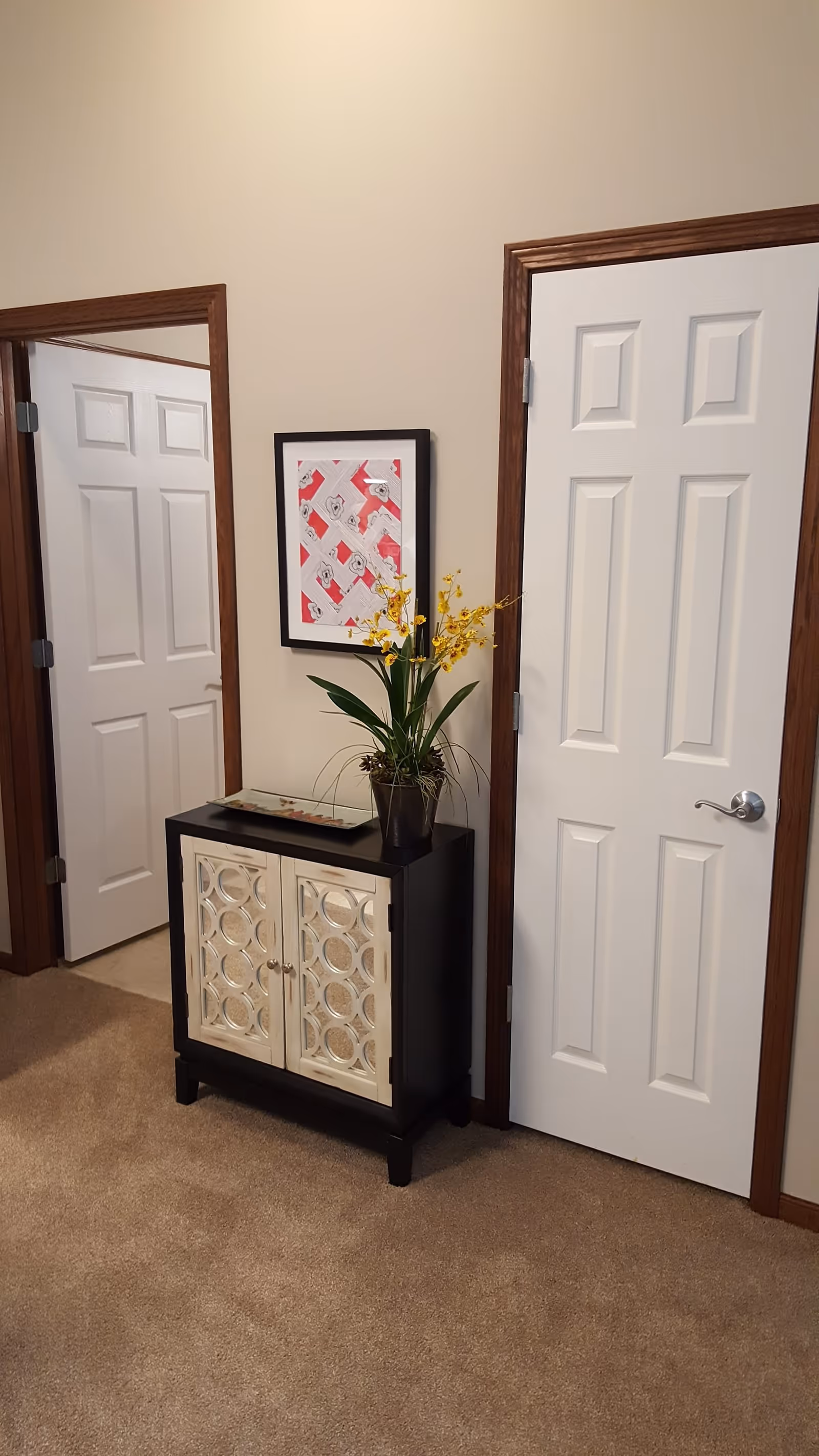 Interior view of a hallway corner in a retirement community with beige carpet and walls. There are two white doors with wooden frames, one slightly open. Between the doors is a small black cabinet with decorative mirrored doors, topped with a potted plant with yellow flowers and a framed abstract artwork hanging above it.
