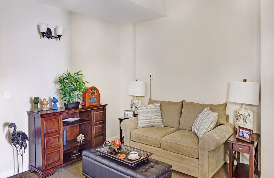 A cozy living room with a beige sofa adorned with two patterned pillows, a dark wooden side table with a decorative lamp and framed photo, a matching wooden cabinet with decorative items and a potted plant, and a black tufted ottoman with a tray holding a cup and snacks.