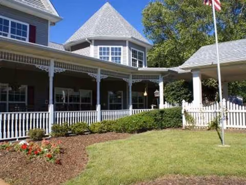 A charming, light-blue senior living building with a wraparound porch and manicured lawn.