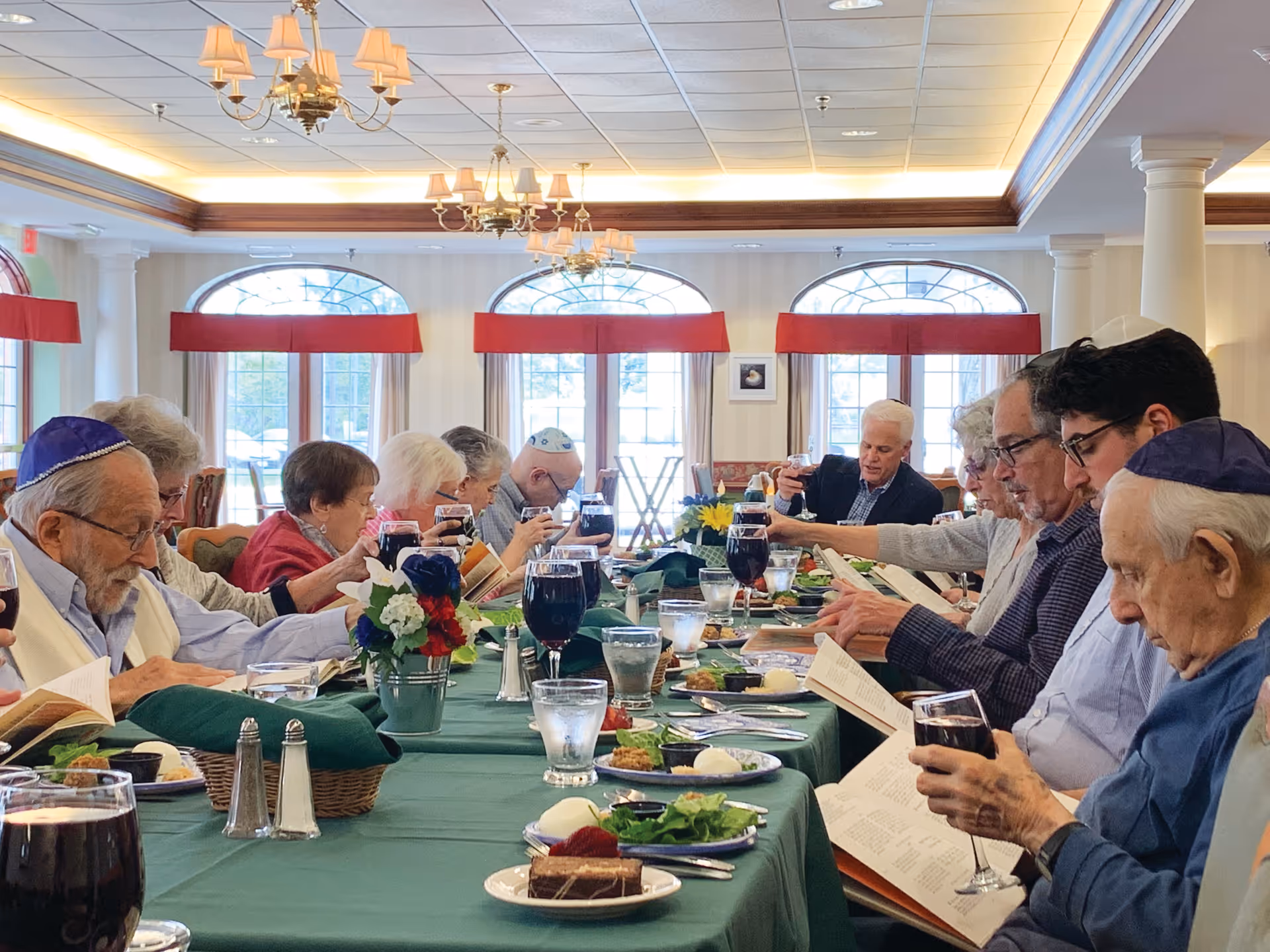 A group of elderly people seated around a long dining table in a well-lit room with large windows and chandeliers. They are holding books and glasses of wine, appearing to be engaged in a communal reading or celebration. The table is set with plates of food, glasses of water, and floral centerpieces.