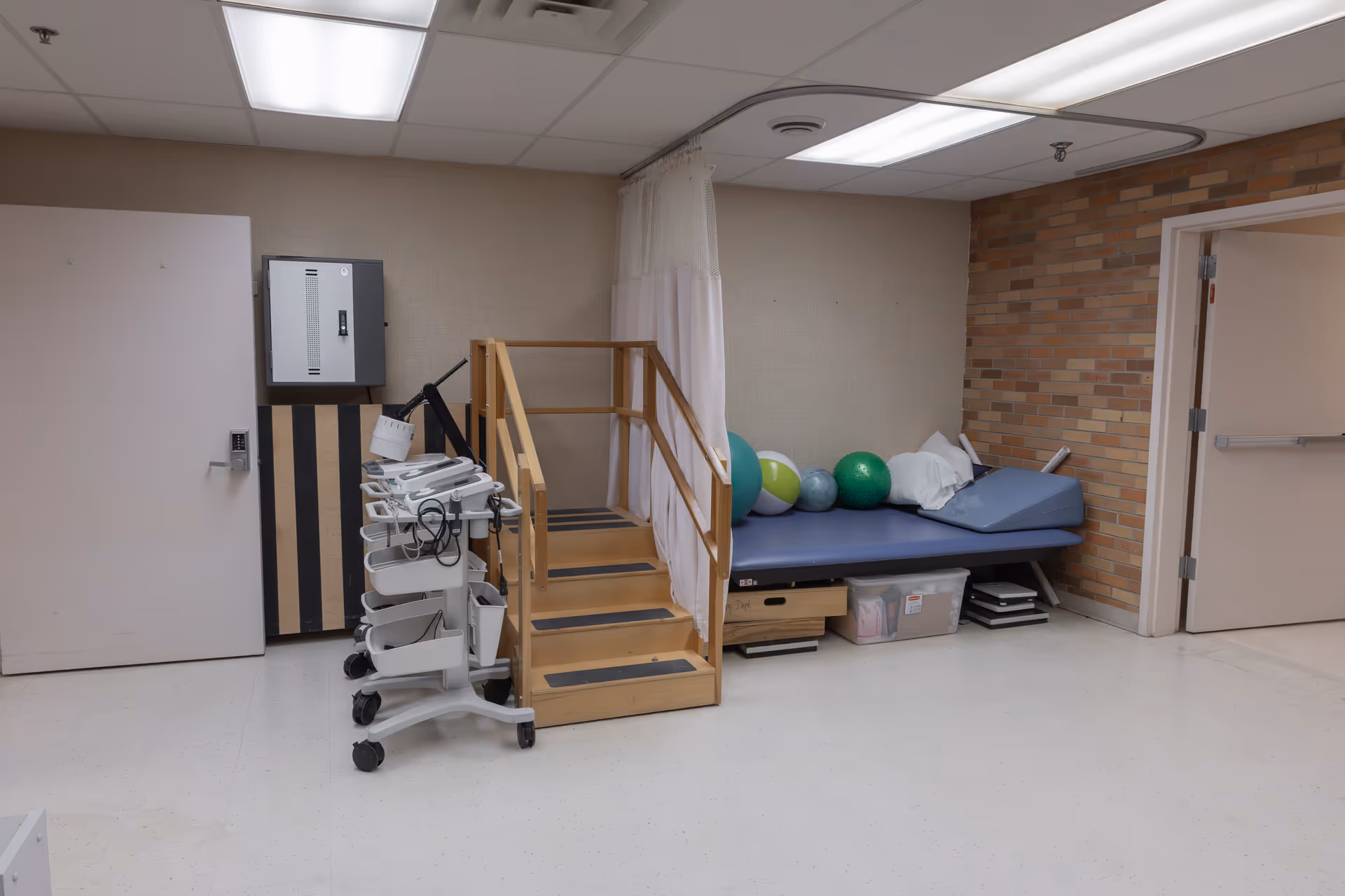 A physical therapy or rehabilitation room with a small wooden staircase, a blue therapy table with pillows and exercise balls, medical equipment on a rolling cart, and a brick accent wall. The room has white tiled floors and fluorescent ceiling lights.