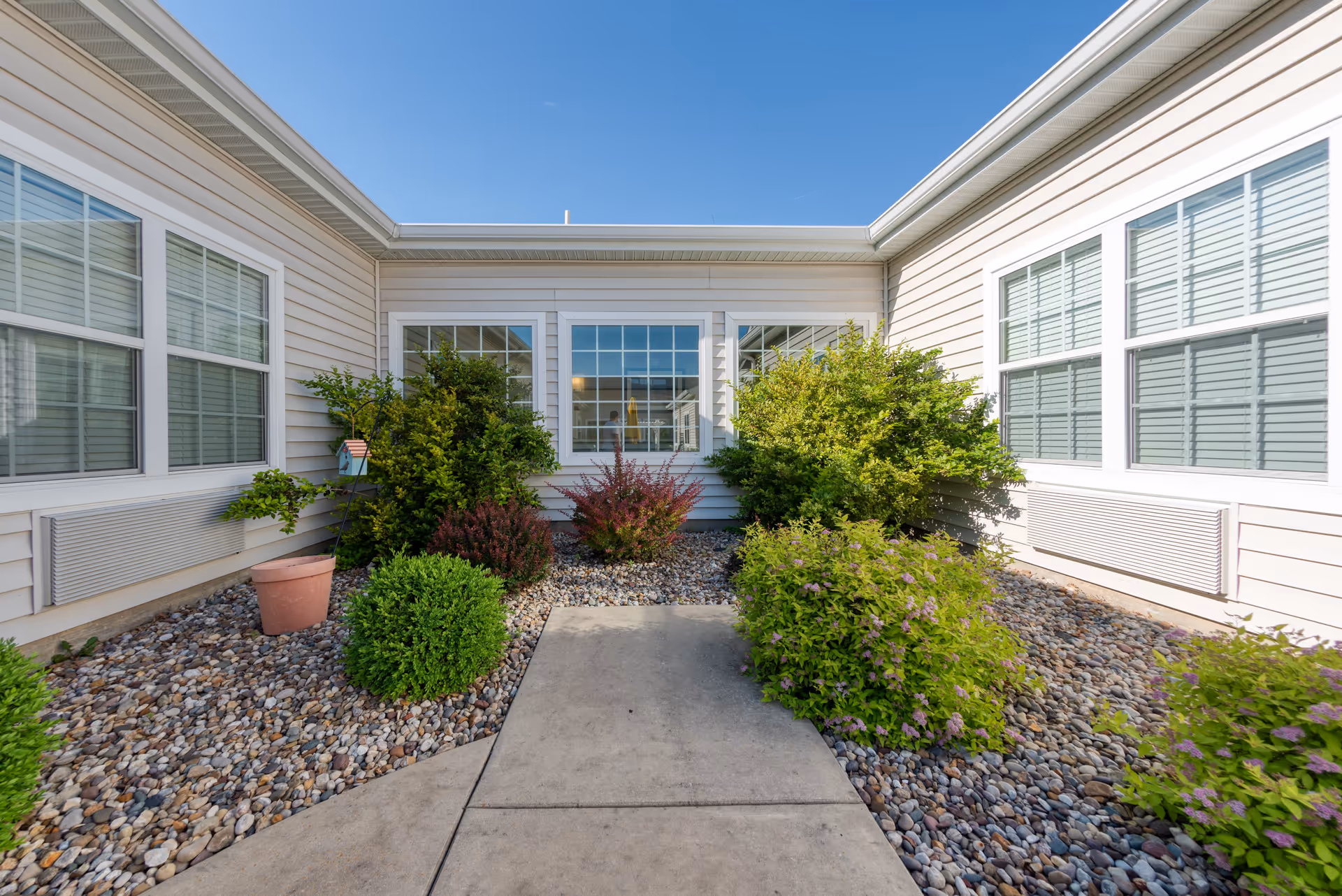 A small outdoor courtyard area with a concrete walkway surrounded by decorative rocks and various green shrubs and plants. The courtyard is enclosed by beige siding walls with multiple white-framed windows, under a clear blue sky.