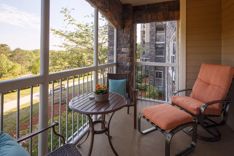 A screened balcony with a small round wooden table, a potted plant with orange flowers, a wicker chair with a blue cushion, and a cushioned lounge chair with an ottoman in orange fabric. The balcony overlooks a green outdoor area with trees and a walking path, and adjacent stone and siding building walls are visible.