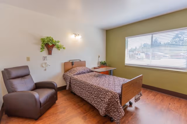 A private nursing facility bedroom with a single hospital bed, recliner chair, nightstand, and a large window with blinds.