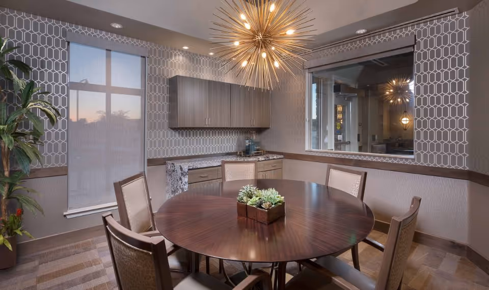 A stylish dining room with a round wooden table surrounded by chairs, a starburst chandelier, patterned wallpaper, cabinets, and a window.