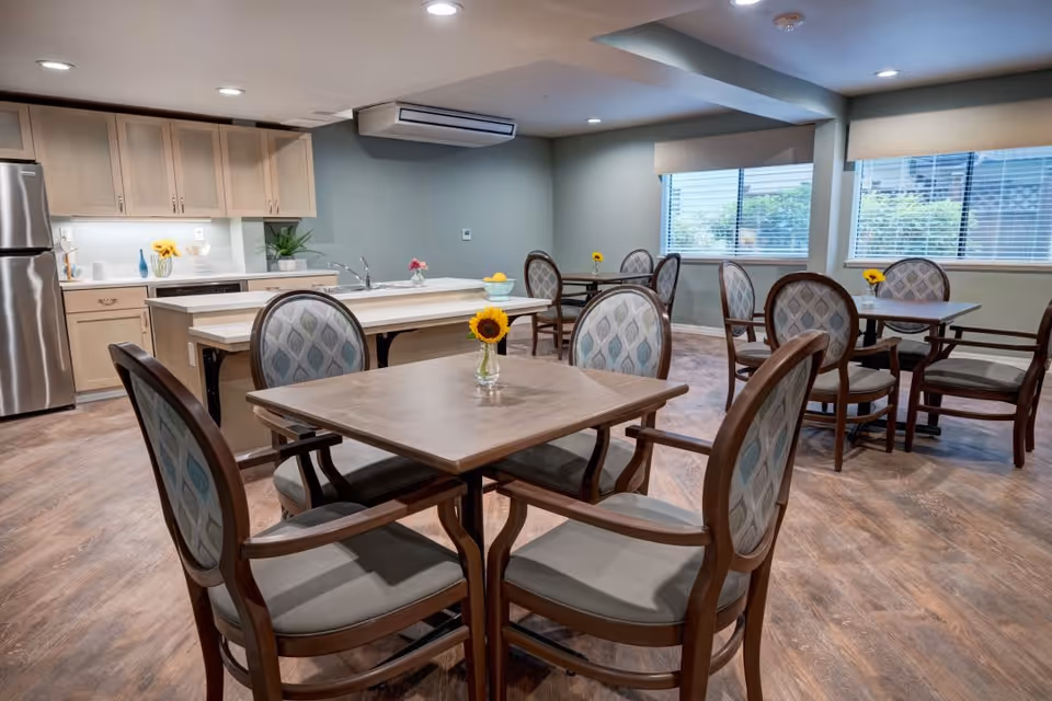A dining area in a senior living facility with several square wooden tables, each surrounded by four upholstered chairs with patterned backs. Each table has a small vase with a sunflower. The room features wood flooring, large windows with blinds, and a kitchen area with light wood cabinets, a stainless steel refrigerator, and a countertop with a sink and decorative items.