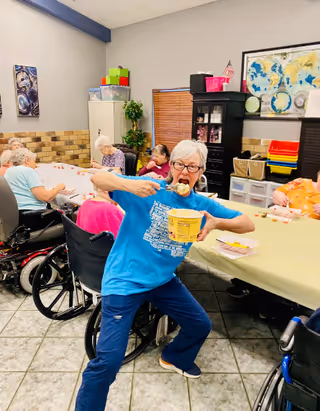 A group of elderly people sitting around tables in a room, some in wheelchairs, engaging in activities. One elderly woman in a blue shirt is standing in the foreground, enthusiastically eating from a large yellow container with a spoon. The room has tiled floors, a map on the wall, storage cabinets, and various colorful bins.