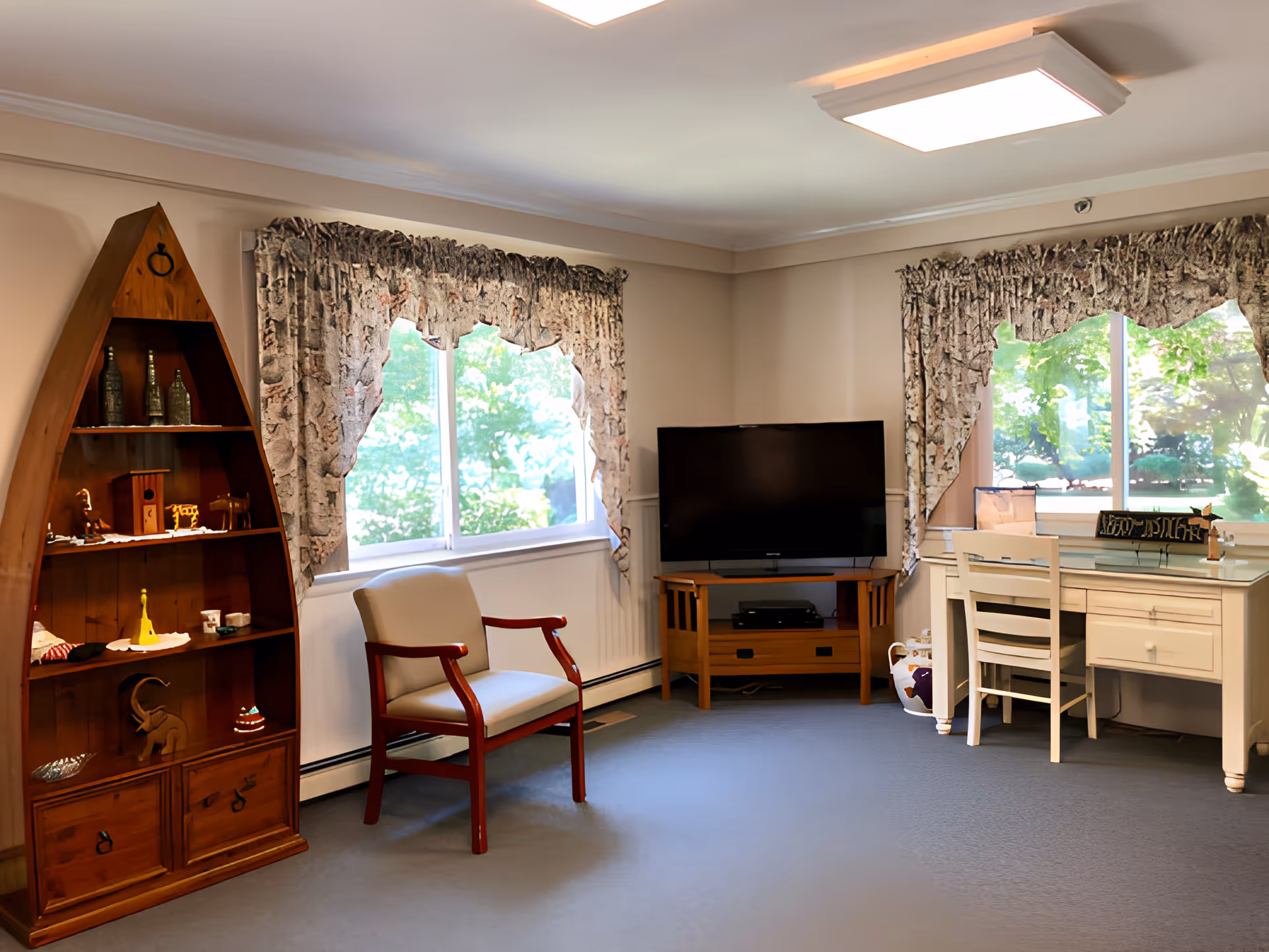 Bright sitting room with a TV on a stand, a boat-shaped wooden shelf, an upholstered chair, and a desk beneath two curtained windows.