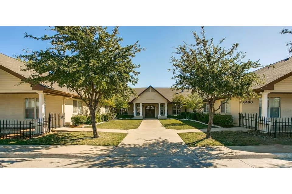 Front exterior view of Avalon Memory Care facility showing a single-story building with a central entrance, two trees, and a paved walkway leading to the door under a clear blue sky.