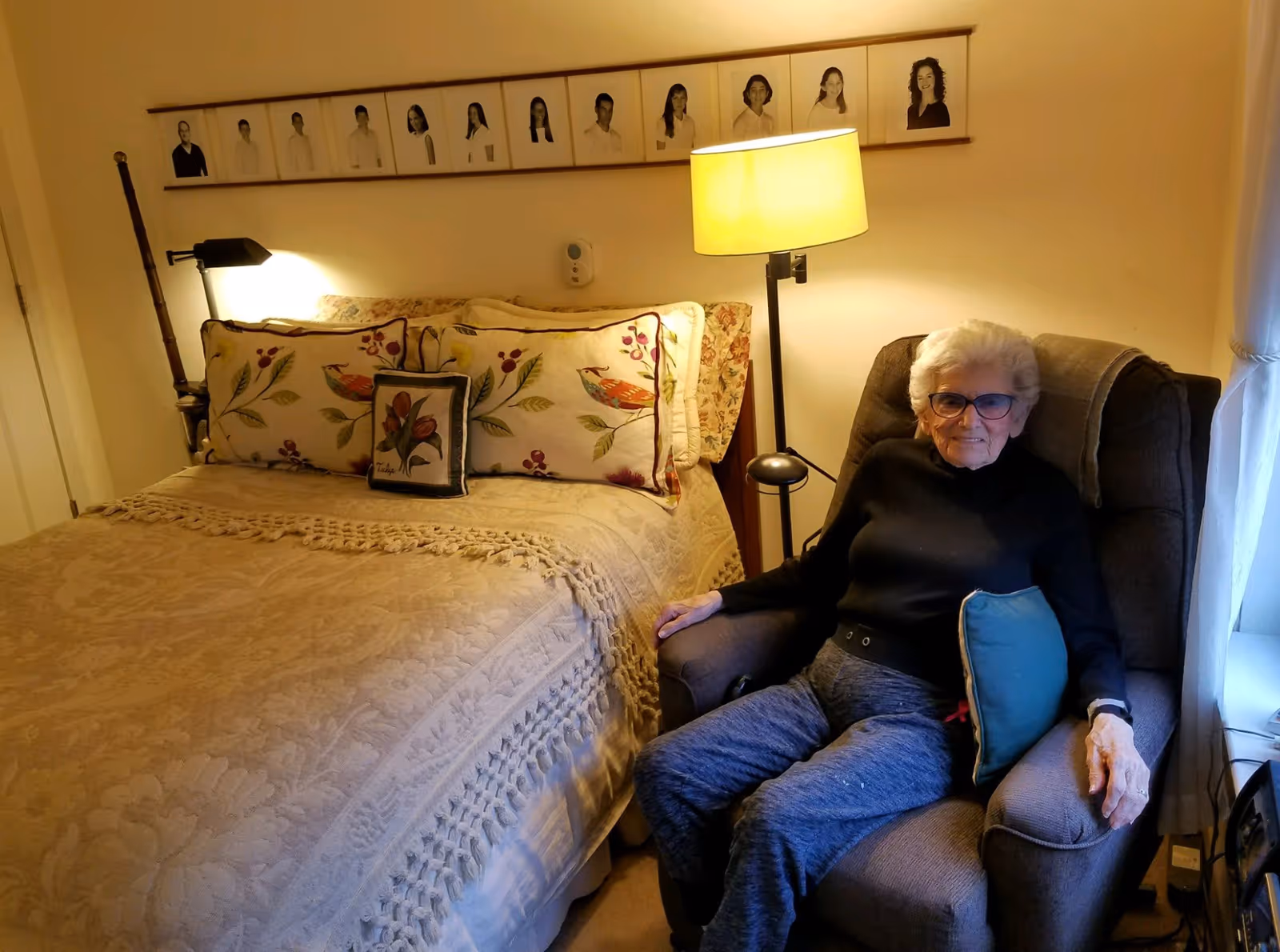 An elderly woman with white hair and glasses sits in a cushioned armchair next to a bed with floral pillows and a beige textured blanket in a warmly lit bedroom. Above the bed is a row of framed black and white portraits on the wall. A floor lamp beside the chair provides additional light.