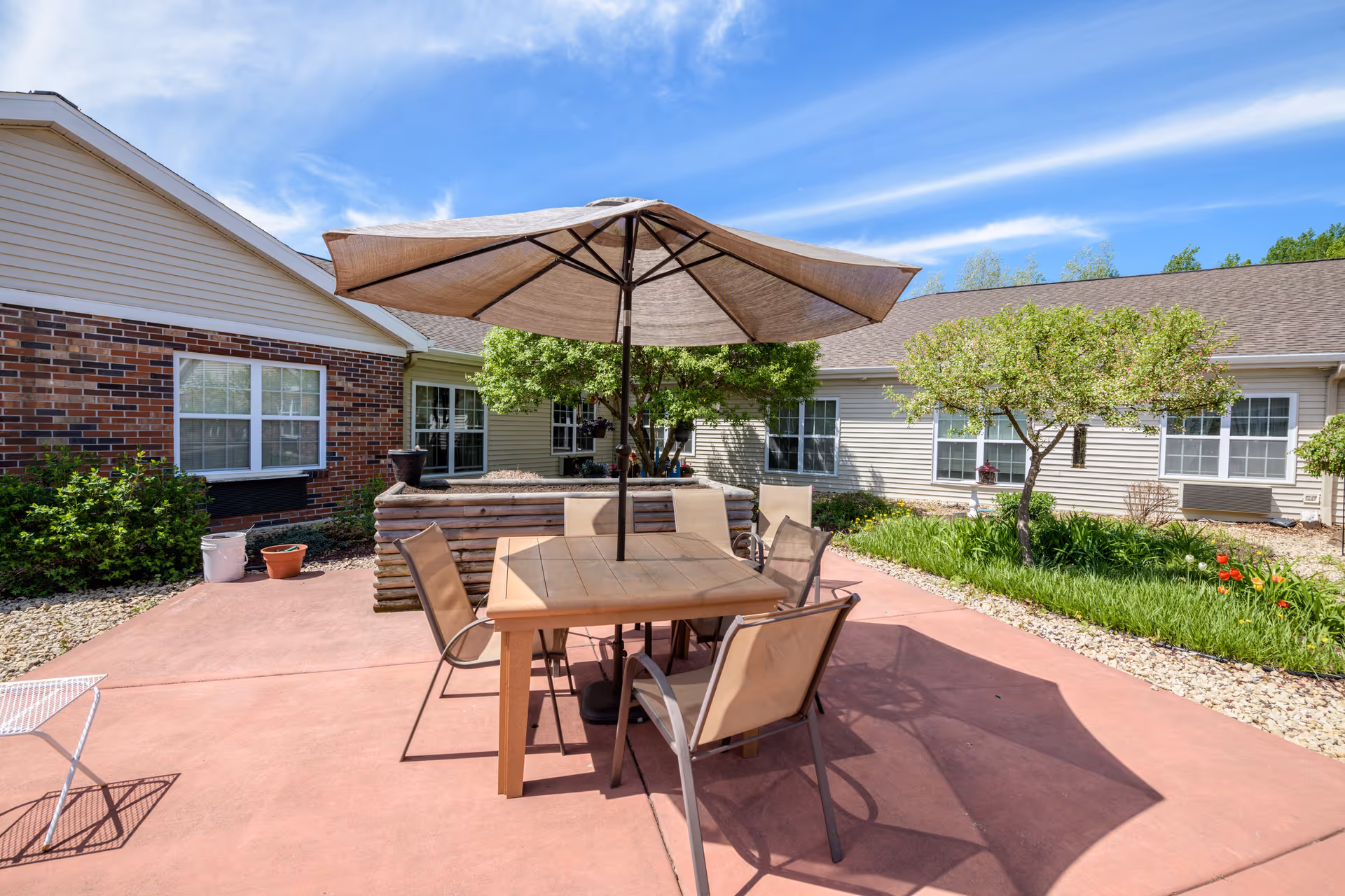 Outdoor patio area at Keystone Bluffs Assisted Living featuring a square wooden table with four chairs and a large beige umbrella providing shade. The patio is surrounded by a garden with green shrubs, small trees, and flowers. The building exterior is visible with brick and siding walls and multiple windows under a blue sky with wispy clouds.