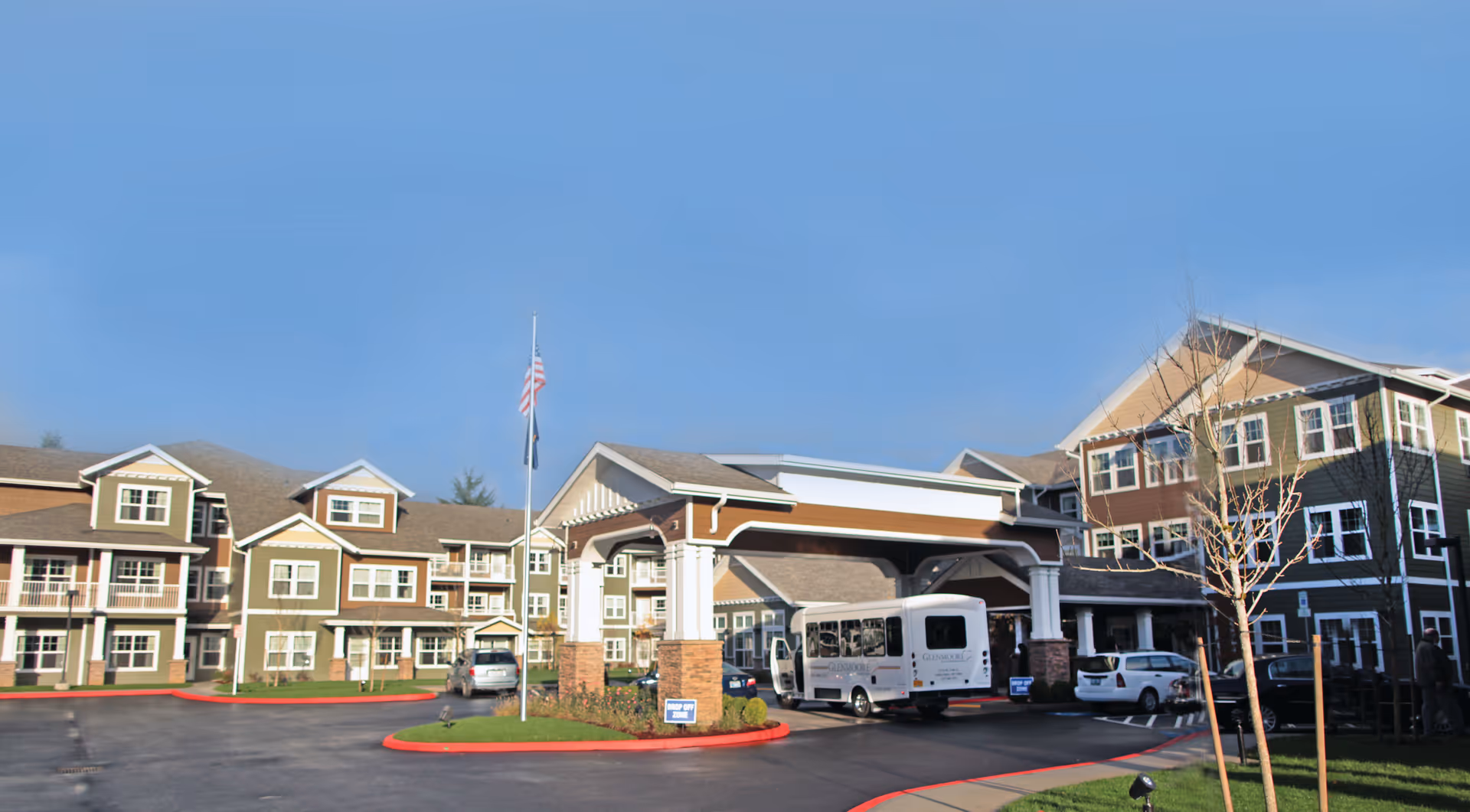 Front exterior of a multi-story senior living building with a covered porte-cochere, parked shuttle bus and cars, and an American flag.