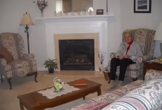 A cozy living room with a fireplace in the center, flanked by two patterned armchairs. An elderly woman is sitting on the right armchair. There is a wooden coffee table with a lace doily and a small flower arrangement in front of the fireplace. A sofa with patterned upholstery is partially visible in the foreground. The room is softly lit with a floor lamp on the left and a table lamp on the right.