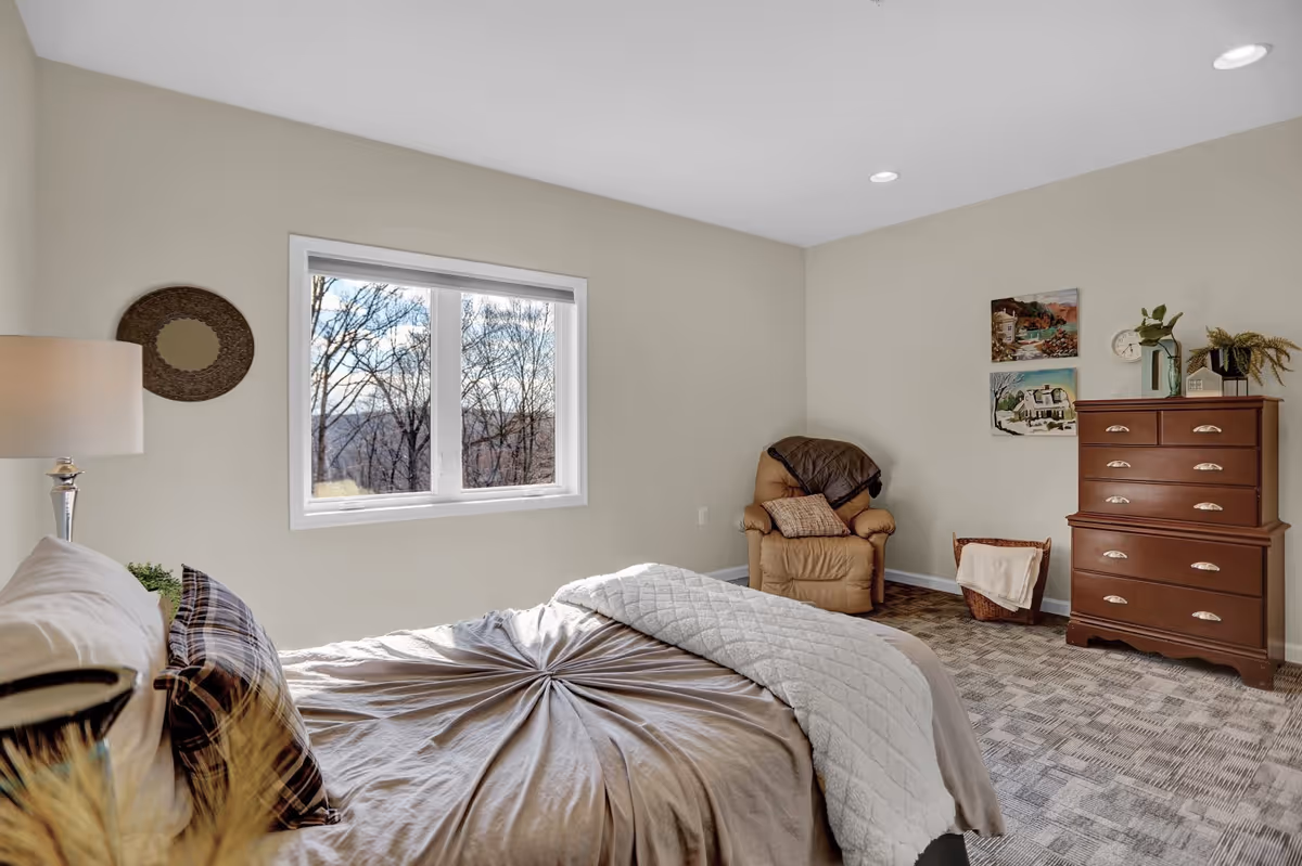 A cozy bedroom with a neatly made bed featuring a beige comforter and plaid pillows. There is a window showing a view of leafless trees outside. The room includes a brown leather armchair with a throw blanket and pillow, a wooden dresser with decorative items and paintings on the wall, and a floor lamp beside the bed.