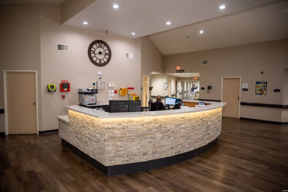 Reception desk area inside a senior living facility with a stone facade and white countertop. Two staff members are seated behind the desk working on computers. The walls are beige with a large round clock mounted above the desk. There are doors and a hallway visible in the background with wood flooring throughout.
