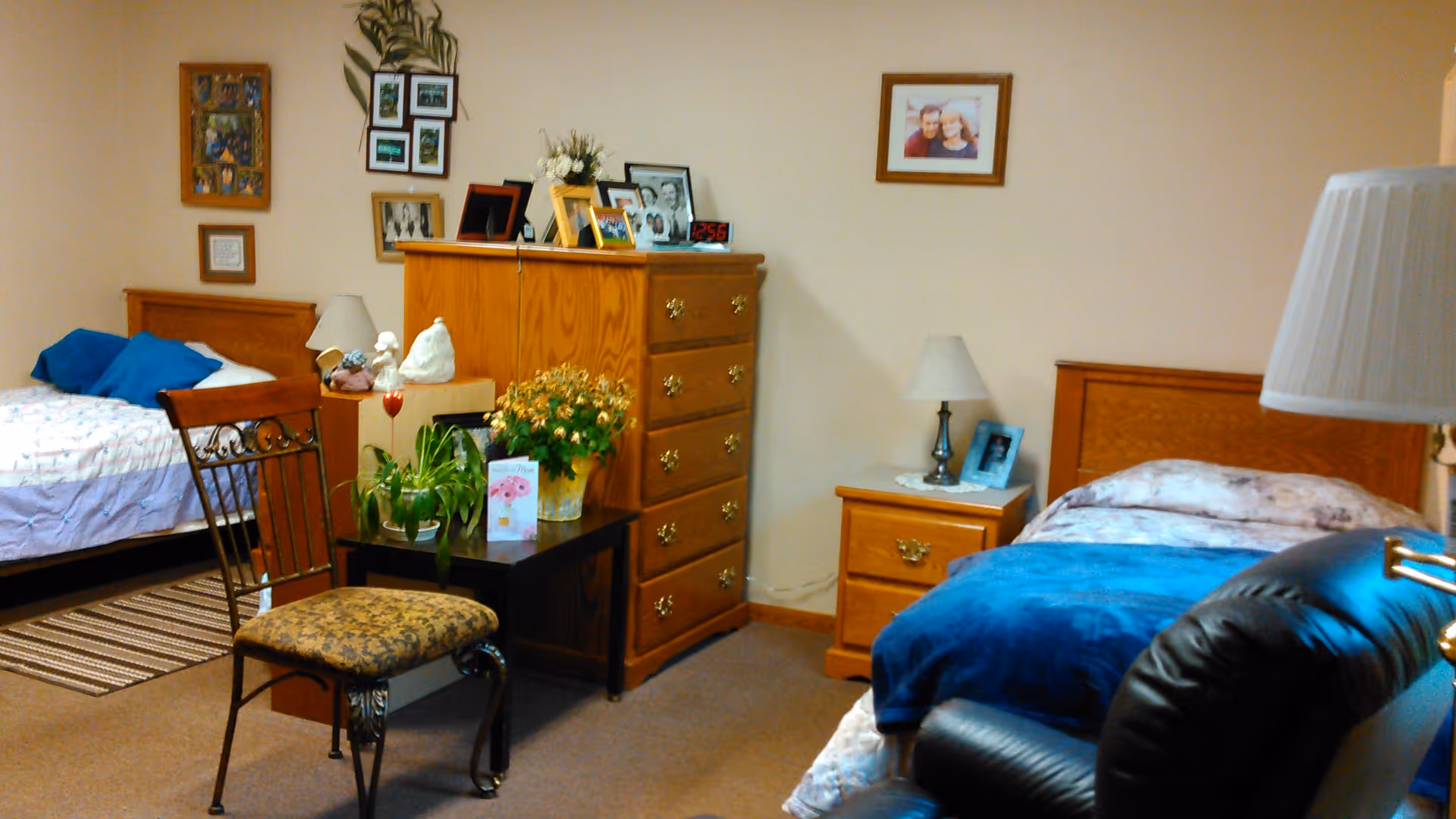A cozy bedroom with two single beds, each with wooden headboards and bedding. Between the beds is a wooden dresser with framed photos and decorative items on top. A small table with plants and a greeting card is in front of the dresser. There is also a wooden nightstand with a lamp and a framed photo. A cushioned chair and a leather recliner are visible in the foreground. The walls are decorated with framed pictures.