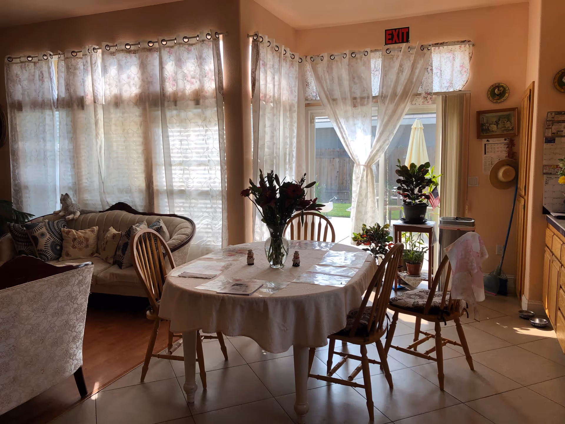 A cozy dining area with a round table covered with a white tablecloth and four wooden chairs around it. A vase with flowers is placed on the table. Behind the table, there is a glass door with sheer white curtains tied in the middle, leading to an outdoor patio with a yellow umbrella. To the left, there is a cushioned sofa with patterned pillows and a cat sitting on the backrest. The room has warm lighting and light-colored walls, with some plants and wall decorations visible.