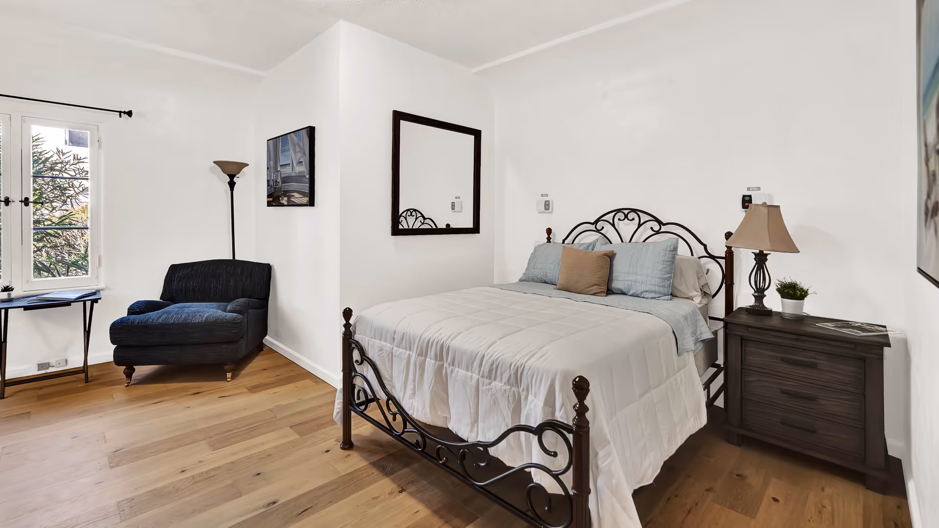 A bright bedroom with a decorative metal bed frame, a neatly made bed with white and blue bedding, a dark wooden nightstand with a lamp and a small plant, a large mirror on the wall, a black armchair near a window with white curtains, and a wooden floor.