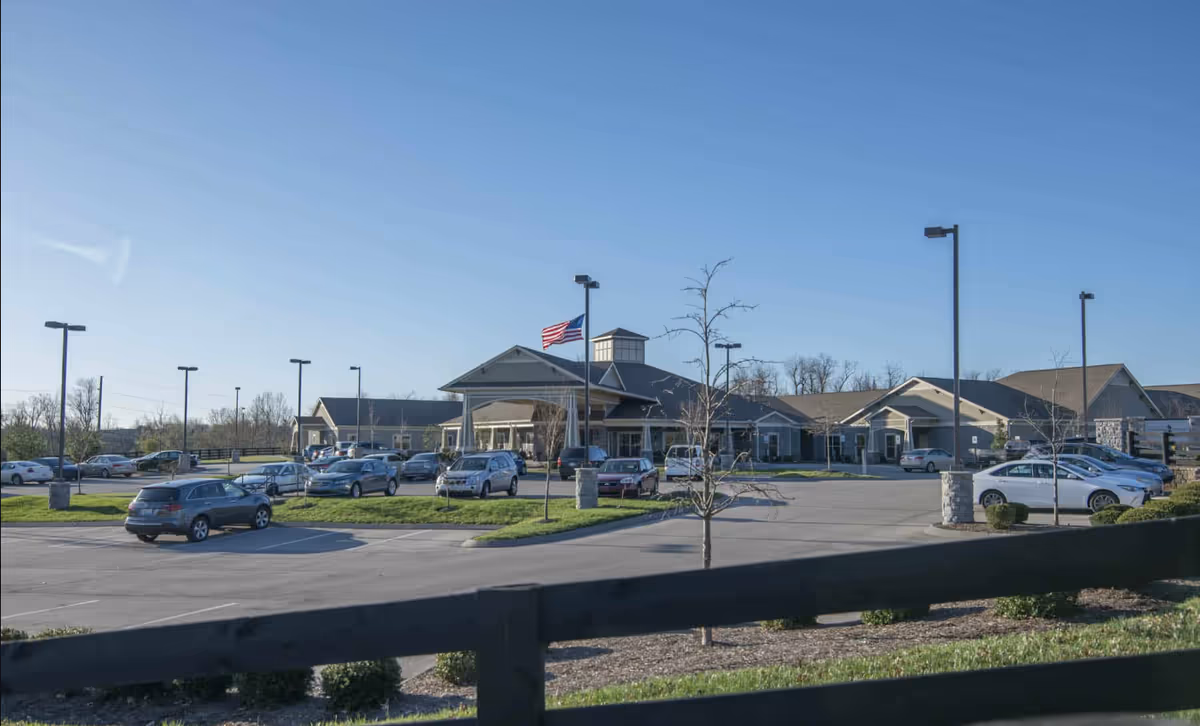 Front exterior view of a senior living building with a parking lot, several parked cars, and an American flag flying near the entrance.