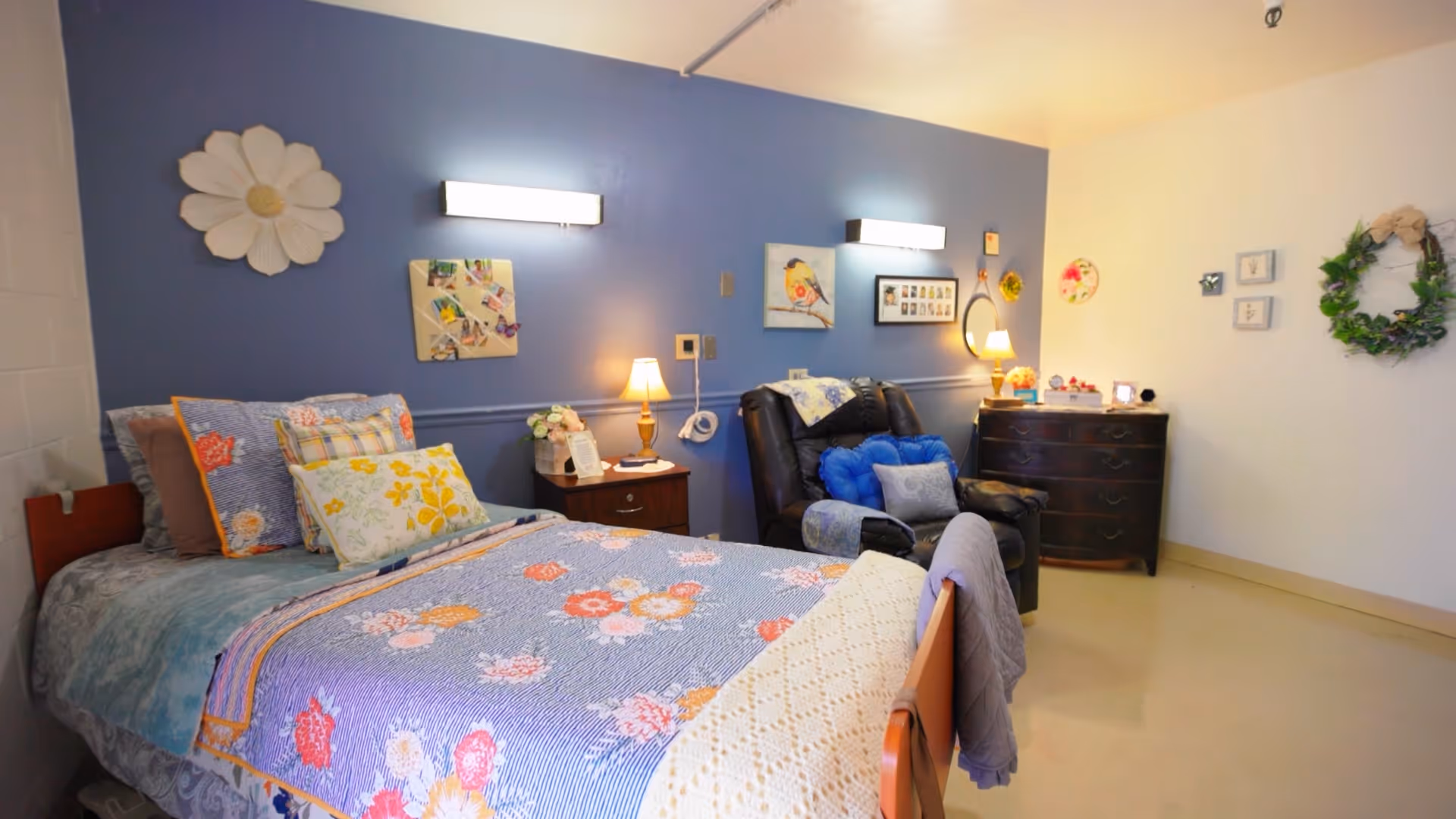 A decorated bedroom with a floral bedspread, bedside lamps, a reclining chair, and a dresser against a blue accent wall.