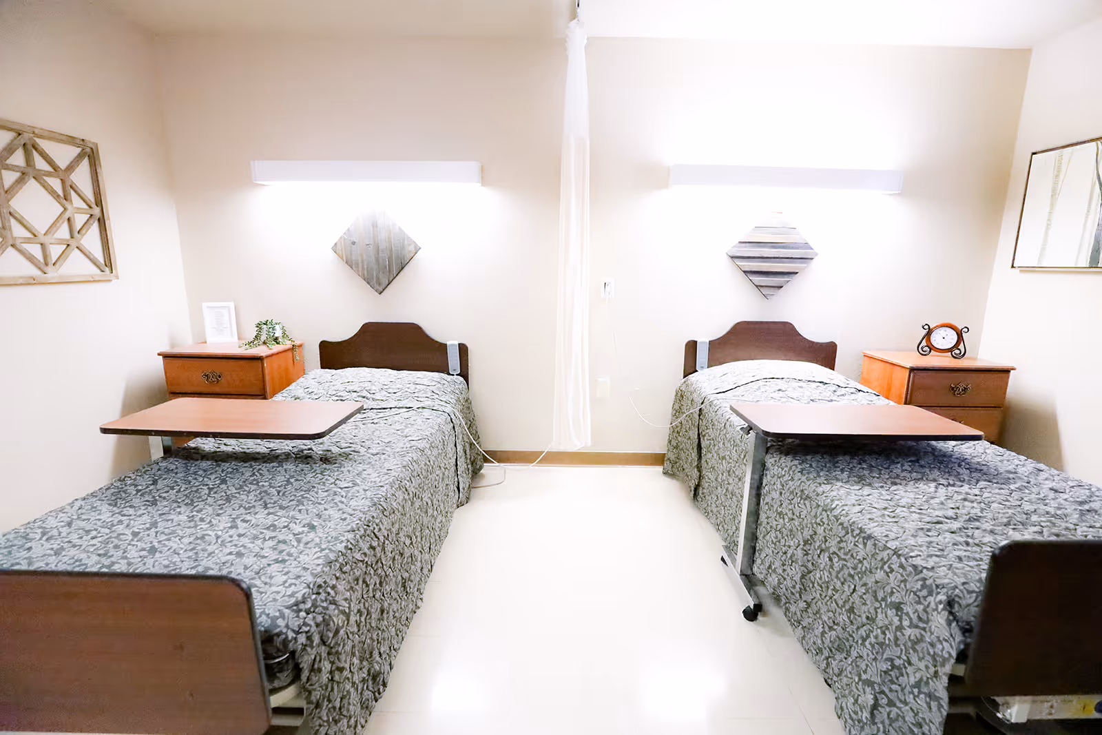 A shared bedroom in a healthcare facility with two single beds separated by a curtain. Each bed has a patterned bedspread, an overbed table, and a wooden nightstand with decorative items. The walls are light-colored with simple wall art above each bed and a mirror on the right wall.