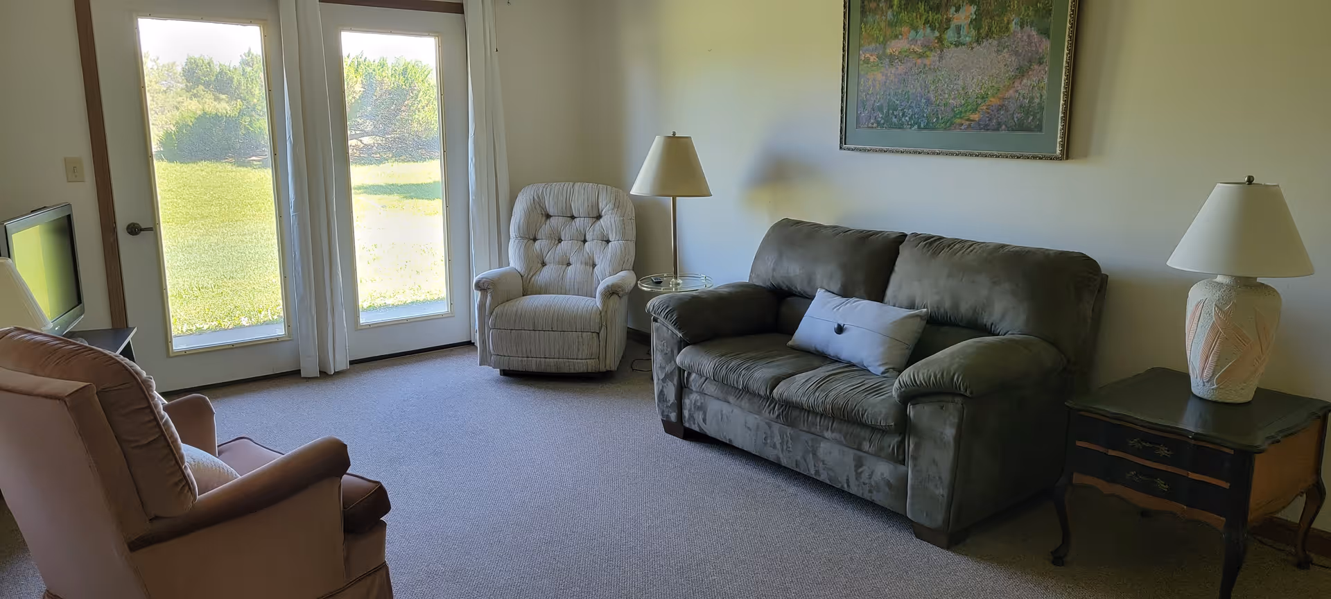 Sunlit living room with a green sofa, two armchairs, side tables with lamps, and double glass doors overlooking a lawn.